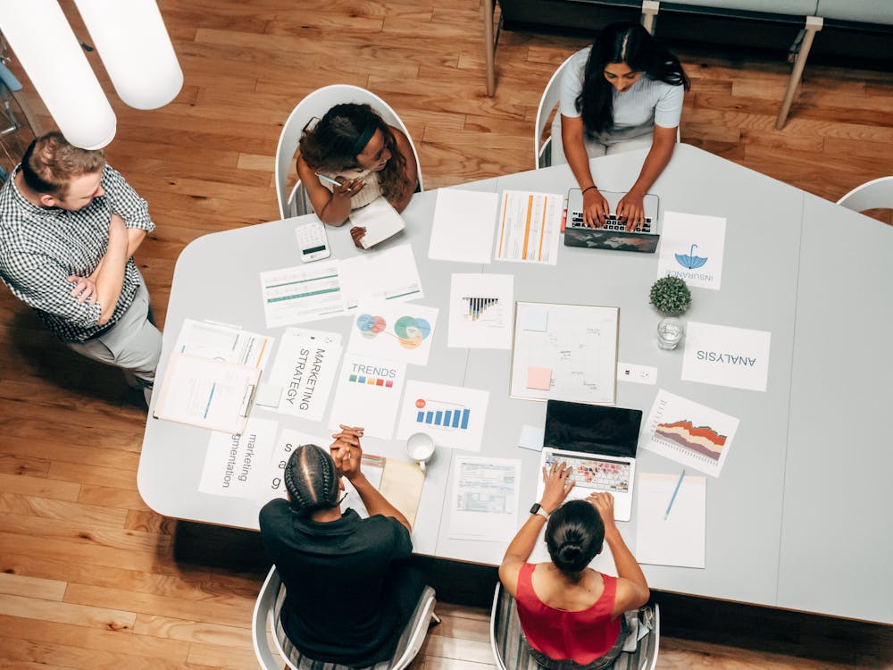 "A diverse group of four people collaborating around a table, working with laptops and reviewing various charts, graphs, and marketing strategy documents. The table is filled with papers displaying data on trends, analysis, and marketing insights. The setting is in a modern office space with wooden flooring."