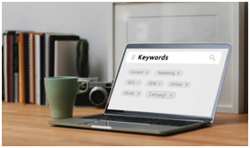 "Open laptop on a desk displaying a keyword search screen with tags related to digital marketing, such as Content, Marketing, SEO, SEM, Online, and Campaign. A green coffee mug and camera are placed beside the laptop, with books in the background."