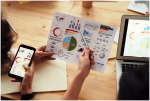 Close-up of a team analyzing data visualizations on printed charts, a smartphone, and a laptop. The charts display colorful graphs, pie charts, and bar graphs showing various statistics and percentages. One person is holding the printed chart and pointing at it with a pen, indicating a discussion or collaborative analysis session at a wooden desk.