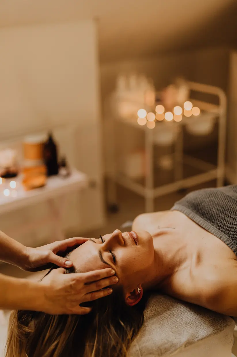 Woman lying on a spa bed with eyes closed while a therapist massages her temples during a facial treatment.